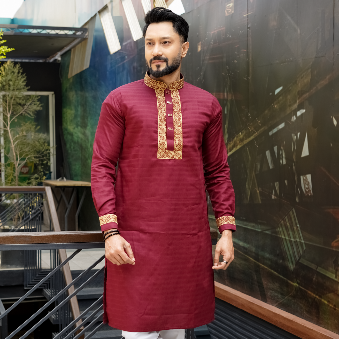 A man wearing a maroon cotton panjabi with gold and green embroidery on the chest and sleeves, paired with white trousers, standing outdoors.