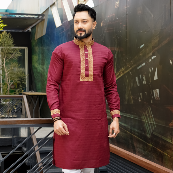 A man wearing a maroon cotton panjabi with gold and green embroidery on the chest and sleeves, paired with white trousers, standing outdoors.